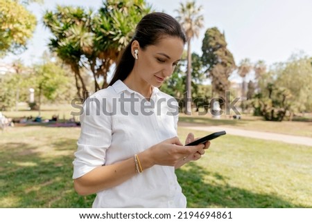 Profile portrait of young brunette woman in casual clothes standing in the park with smartphone and listening music in wireless headphones on park background 