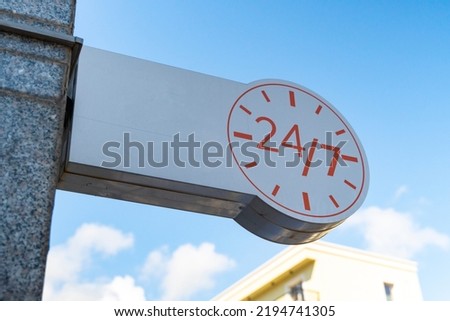 A white banner with the image of a clock signifying the round-the-clock operation of the institution against the background of a blue sky. twenty-four by seven round-the-clock work signage
