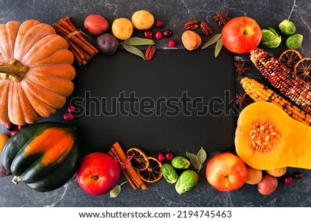Fall food frame of pumpkins, apples, squash and an assortment of vegetables surrounding a blank slate serving board. Top down view on a dark stone background with copy space.