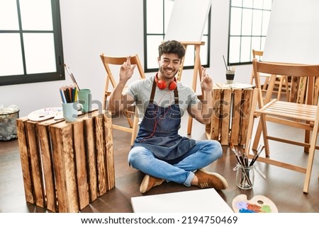 Young hispanic man sitting at art studio gesturing finger crossed smiling with hope and eyes closed. luck and superstitious concept. 