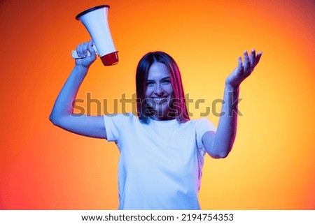 Shouting at megaphone. Young emotional girl, student in white t-shirt isolated on orange color background in neon light. Concept of info, propaganda, news