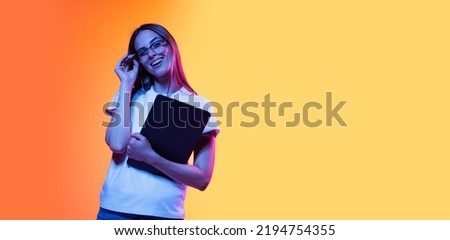 Enjoying student life. Portrait of young smiling girl in white t-shirt with laptop isolated on orange color background in neon light. Concept of beauty, art, fashion, education