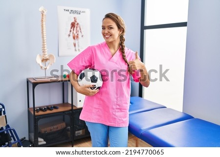 Young hispanic woman working at physiotherapy clinic holding football ball smiling happy and positive, thumb up doing excellent and approval sign 