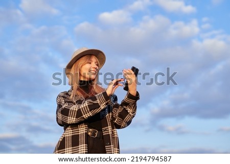 Attractive hipster girl making online shopping in background of blue cloudy sky. Young caucasian blonde female in hat holding smartphone and credit card. High quality image