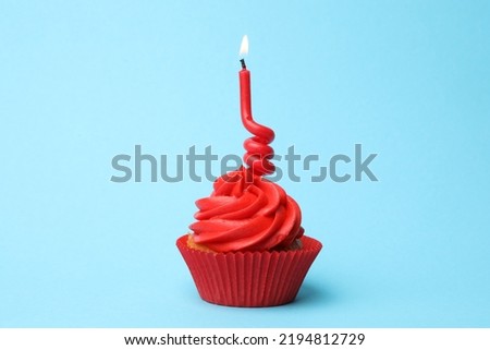Delicious birthday cupcake with red cream and burning candle on light blue background