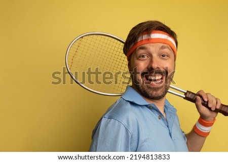 Studio portrait of a 40 year old attractive caucasian man wearing vintage clothing and holding a vintage tennis racquet. The studio background is yellow.
