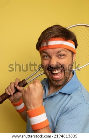 Studio portrait of a 40 year old attractive caucasian man wearing vintage clothing and holding a vintage tennis racquet. The studio background is yellow.