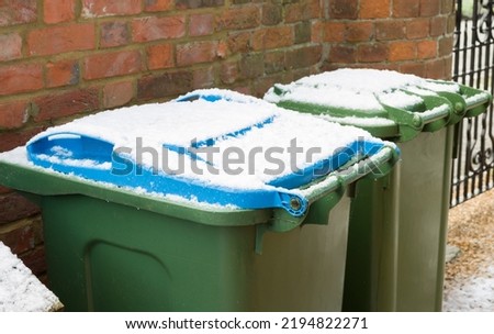 Green and blue wheelie bins in a garden in winter, UK. Bin lids covered in snow. Royalty-Free Stock Photo #2194822271