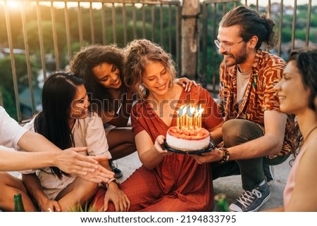 A group of hippie multicultural friends surprises a birthday girl with a cake at a rooftop party. A man holds a cake and gives it while the rest of the friends wish all the best.
