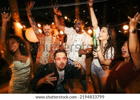 A group of multiracial friends is at the open-air rooftop club at night and having a good time. They are dancing, drinking beer, and celebrating summer. There is confetti everywhere. Royalty-Free Stock Photo #2194833799