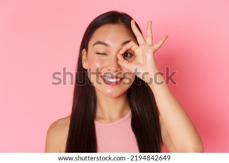 Beauty, fashion and lifestyle concept. Portrait of kawaii attractive asian girl showing okay gesture over eye and winking carefree at camera, smiling pleased, guarantee quality, recommend place