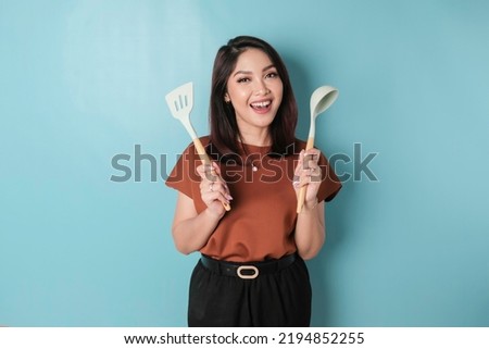 Excited Asian woman holding cooking ware and smiling, isolated by blue background