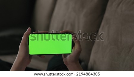 Young woman lying on a couch and using smartphone with horizontal green screen, wide photo