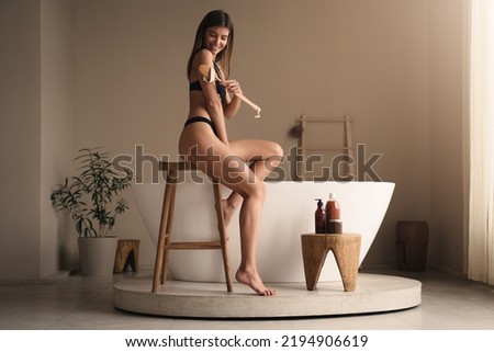 Woman peeling body with dry brush doing lymphatic drainage massage sitting on wooden stool in bikini over modern ceramic bathtub, having beauty and spa procedures in bathroom at home