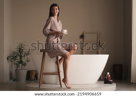 Portrait of happy relaxed female in silk robe drinking herbal tea after beauty and spa procedures, sitting on high wooden stool with white ceramic bathtub on background in fashionable bathroom Royalty-Free Stock Photo #2194906659