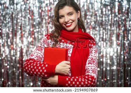 Portrait of smiling woman in knitted winter clothes holding christmas present box on tinsel background. Merry xmas. Happy new year