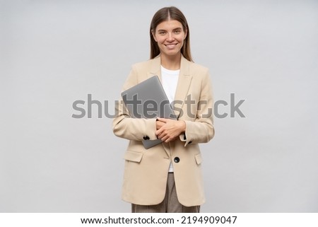 Portrait of pretty young woman teacher with laptop in hands greeting her students, ready to start lesson, dressed in beige jacket isolated on gray copy space background