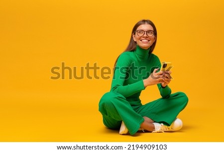 Dreamy student girl in stylish green clothes sitting with crossed legs on floor using smartphone, having rest on yellow copy space background holding phone in hands, looking up aside Royalty-Free Stock Photo #2194909103