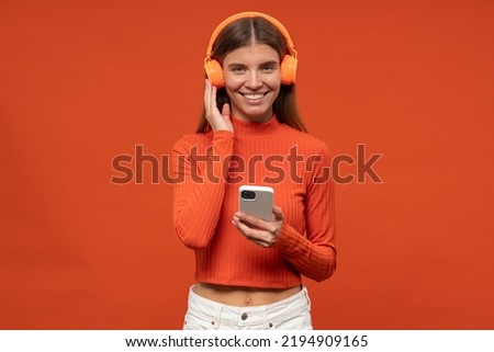 Pretty millennial music fan in stylish clothes and wireless headphones holding smartphone listening to classic music on orange studio background smiling at camera
