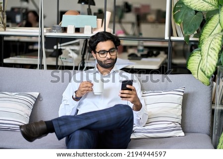 Indian professional business man executive holding smartphone using mobile phone reading news, checking financial market or planner applications sitting on sofa drinking coffee working in office.