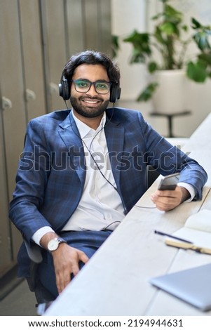 Young happy indian business man employee executive manager wearing headphones sitting at workplace using mobile phone looking at camera in office. Audio training podcasts course ads. Vertical