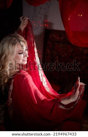 A beautiful European girl looking like an Arab woman in red room in a harem. Photo shoot of an oriental style odalisque. A model poses in a sari as an indian woman in india