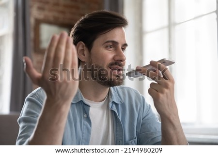 Positive engaged millennial cellphone user man talking on speaker on cellphone, sitting at table at home office workplace, recording audio message, giving voice command. Communication concept