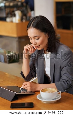 Young professional Asian business woman wearing suit using apps on digital tablet sitting at table remote working or elearning online, holding stylus writing e signature signing electronic document.