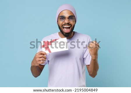 Young happy unshaven student black african man in violet t-shirt hat glasses hold gift voucher flyer mock up do winner gesture clench fist clebrate isolated on pastel blue background studio portrait