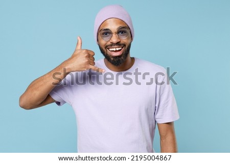 Young smiling happy positive friendly unshaven black dark-skinned african man in violet t-shirt hat glasses do phone gesture like says call me back isolated on pastel blue background studio portrait