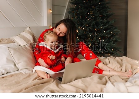 Mom and daughter in red pajamas are looking at a laptop, preparing for the Christmas holiday and ordering gifts online using technology while lying on the bed at home in winter. Selective focus