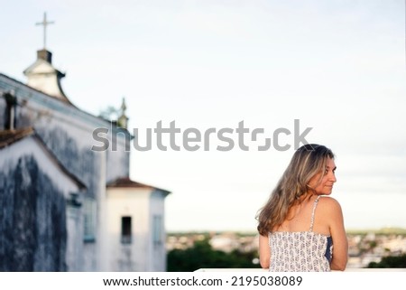 A woman on the porch with her back to the camera against the sky and church in the background. City of Valença, Bahia, Brazil.