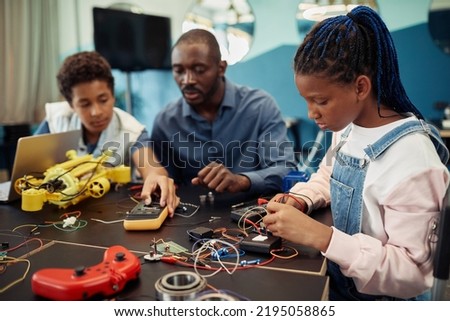 Side view portrait of young black girl building robots with male teacher in background during engineering class at school Royalty-Free Stock Photo #2195058865