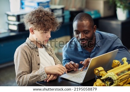 Portrait of black male teacher helping young boy building robot during engineering class in school and using laptop for programming together Royalty-Free Stock Photo #2195058879
