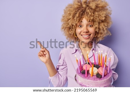 Horizontal shot of positive curly haired woman holds festive cake with burning candles points away on blank space for your advertising content dressed in shirt isolated over purple background.