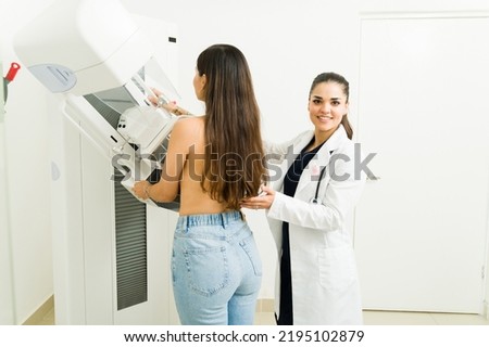 Happy female doctor or radiologist on a lab coat smiling while helping a young woman during a mammogram medical exam