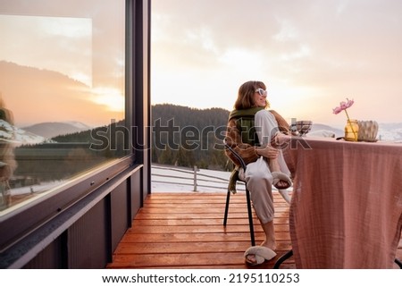 Woman sitting on terrace with great view on the mountains in winter, enjoying drink and beautiful landscape during sunrise. Concept of rest and escape to nature