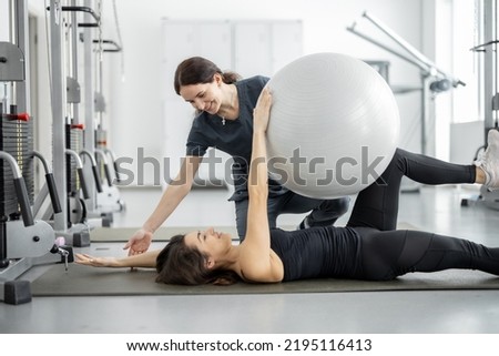 Woman doing exercises with fitness ball with rehabilitation specialist at the gym. Concept of physical therapy for health and recovery. Idea of recovery after pregnancy Royalty-Free Stock Photo #2195116413