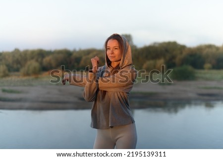attractive young caucasian girl performing stretching at dawn at bank of river, dressed in grey hoodie and leggins, morning relaxation, sporty lifestyle