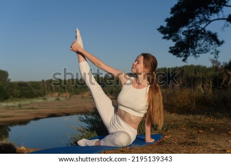 attractive young caucasian girl doing stretching at dawn at bank of river, sitting on blue gym mat, dressed in white top and leggins, morning relaxation, sporty lifestyle