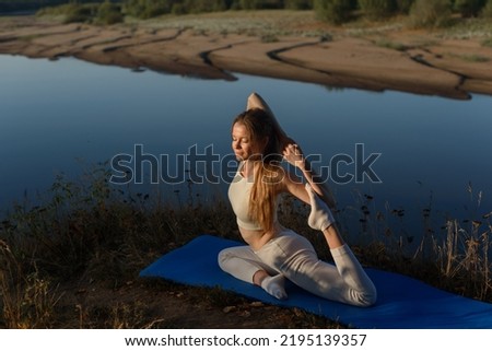 attractive young caucasian girl doing stretching at dawn at bank of river, sitting on blue gym mat, dressed in white top and leggins, morning relaxation, sporty lifestyle