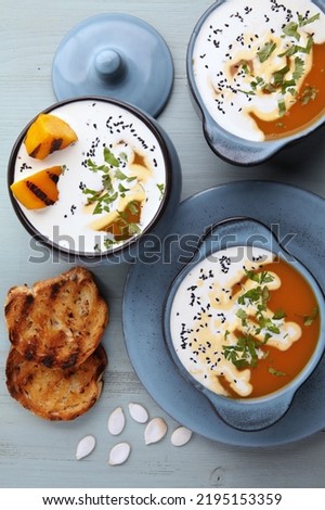 Hot soup puree with cream, black cumin seeds, herbs, pumpkin seeds and toasted bread on a blue wooden table. Thick soup in bowl and pot
