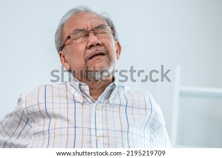 An agonizing senior man in need of assistance while sitting on his sofa at home, suffering from back pain. Senior care, nursing home for pensioners, deteriorating health of old age.