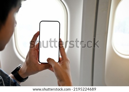 Young Asian male traveler or passenger using his smartphone to taking a picture of beautiful sky during the flight. close-up image, phone white screen mockup.