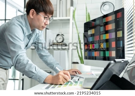 Male graphic designer is standing to choosing color swatch samples on multiple screens and working graphic design with technology while sketching on tablet digital in modern office.