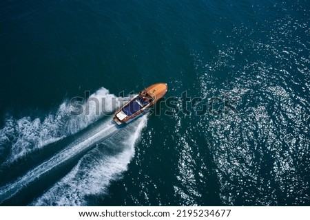 An expensive wooden boat is an average movement on the water. Classic wooden boat with motor moving on blue water aerial view. Italian wooden boat fast moving diagonal top view. Royalty-Free Stock Photo #2195234677