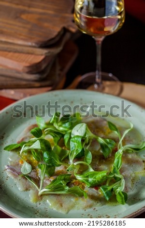 Ocean bass carpaccio with wine vinegar, microgreen leaves. The food lies on a ceramic green plate. The plate stands on a red, wooden background, boards lie next to it and a glass of white wine stands.