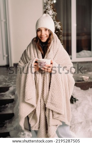 Full-length photo of a beautiful happy little girl wearing white hat and blanket holding a cup of hot tea outside on winter evening.