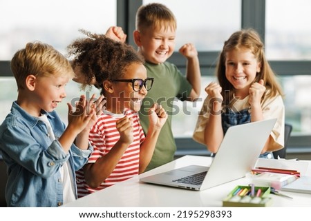 Positive kids pupils  schoolchildren    happy smiling and   looking at laptop screen during celebrate together  successful completion of collective school work in a light classroom   Royalty-Free Stock Photo #2195298393