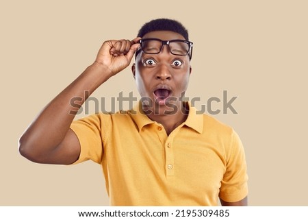 Young African American man in polo shirt opens his mouth, lifts glasses, looks at something with surprised shocked astonished face expression and says Wow, just look at that. Studio head shot